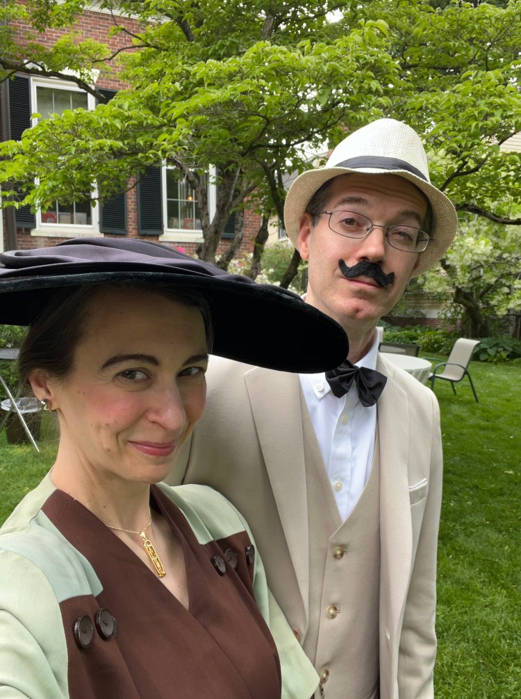 Julie and Bryan Butters dressed as Agatha Christie and Hercule Poirot respectively, in a vintage brown and mint-green dress and wide-brimmed black hat and beige suit with matching hat, glasses, black moustache and black bow tie, against the backdrop of the rear of the Salem Athenaeum in Salem, MA